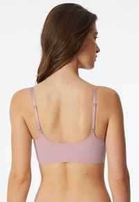 Woman seen from the back wearing a mauve bra with thin adjustable straps against a plain light background.