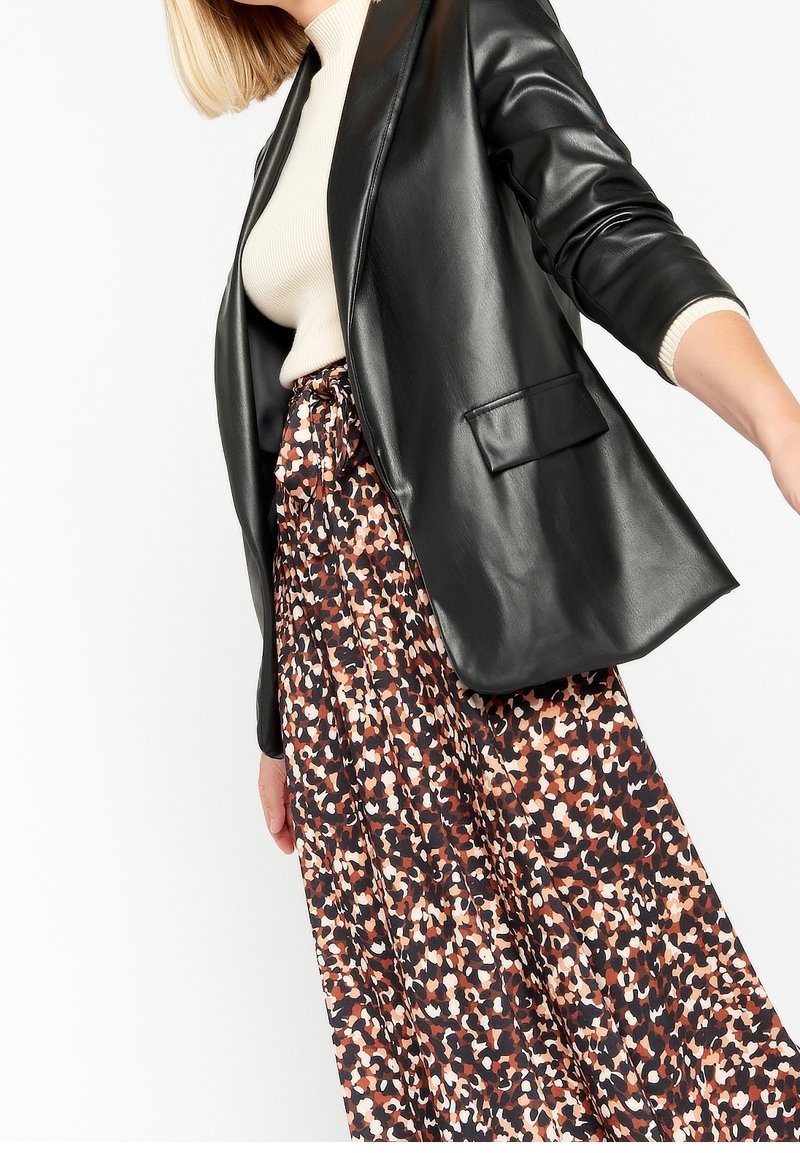 Femme portant un haut côtelé beige, un blazer noir en similicuir et une jupe taille haute à motif abstrait multicolore.