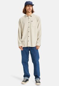 Beige, button-up shirt with long sleeves, one chest pocket, paired with blue jeans and black canvas shoes. Dark blue cap completes the outfit.