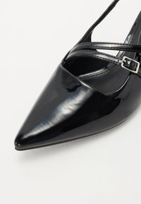 Crne lakirane cipele na visoku petu s uskim vrhom, s dizajnom križnog remena i detaljem od srebrne kopče. Glatka, sjajna tekstura i elegantan oblik.