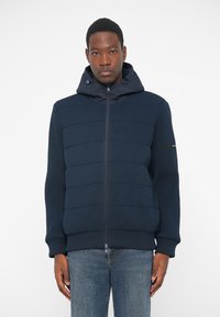 Μπλέ φούτερ με φερμουάρ και ραμμένο υλικό με ραβδώσεις και αφράτες επενδύσεις. Διαθέτει κουκούλα και ελαστικέ�ς μανσέτες, φορεμένο με ανοιχτόχρωμα τζιν.