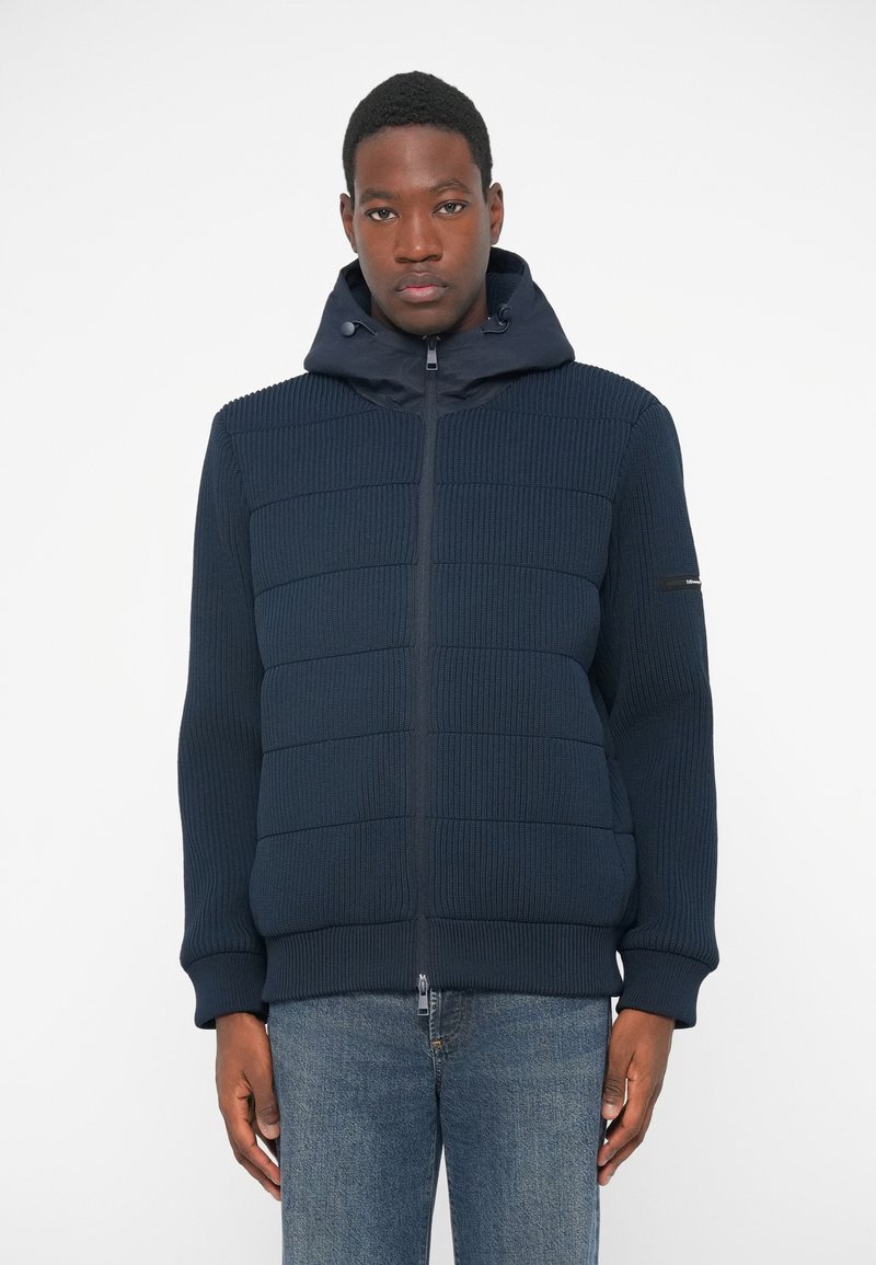 Μπλέ φούτερ με φερμουάρ και ραμμένο υλικό με ραβδώσεις και αφράτες επενδύσεις. Διαθέτει κουκούλα και ελαστικές μανσέτες, φορεμένο με ανοιχτόχρωμα τζιν.