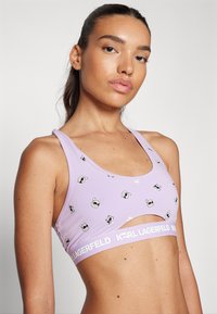 Bra sportivo viola con un design a taglio, caratterizzato da motivi a cartone animato giocosi e una banda con logo bianco nella parte inferiore. Tessuto morbido e elasticizzato.