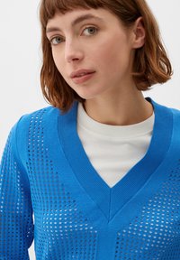 Blauer Strickpullover mit V-Ausschnitt und offener Lochmuster; über einem weißen Hemd getragen. Glatte Textur und leichtes Design.