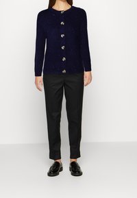 Granatowy sweter rozpinany z prążkowanej dzianiny z okrągłym dekoltem i sześcioma dużymi guzikami, zestawiony z czarnymi, dopasowanymi spodniami i błyszczącymi czarnymi mokasynami.