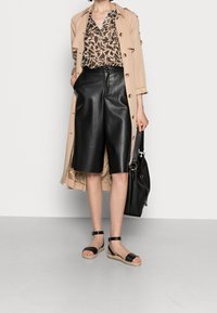 Trench beige, blusa marrón estampada, pantalones cortos de cuero negros, sandalias espadrilles negras y un bolso negro. Texturas suaves y colores neutros.
