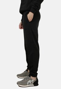 Pantaloni della tuta neri realizzati in tessuto morbido con un design affusolato, dotati di tasche e polsini elastici, indossati con scarpe sportive grigie.