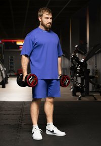 Maglietta atletica blu e pantaloncini, con in mano manubri neri e rossi. Sneakers bianche con accenti neri, in piedi su un pavimento di palestra.