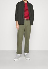 Mænds outfit med en sort skjorte med knapper, en rødstribet t-shirt, olivenfarvede bukser og grå slip-on sko med hvide såler.