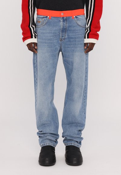 Jeans in denim azzurro chiaro con una vestibilità rilassata, dotati di una cintura arancione, cinque tasche e orli risvoltati. Scarpe nere completano l'outfit.