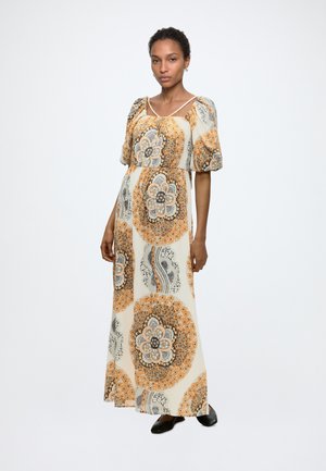 Frau trägt ein beiges Maxikleid mit orange-schwarzen kreisförmigen Blumenmustern, Puffärmeln und schwarzen flachen Schuhen, steht vor weißem Hintergrund.