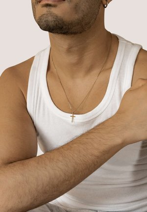 Mann mit gestutztem Bart und Ohrring, der ein weißes Tanktop und eine goldene Kreuzkette trägt, mit über die Brust verschränkten Armen.