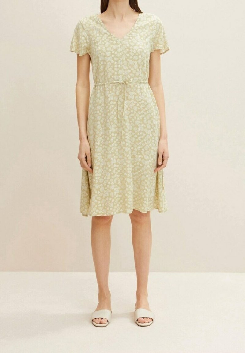 Femme portant une robe jaune clair à fleurs, longueur genou, manches courtes avec ceinture à nouer, assortie à des sandales blanches à enfiler.