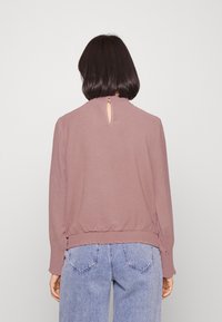 Suéter rosa empolvado con escote redondo, tejido texturizado, detalle en la espalda con forma de llave y puños fruncidos. Combinado con jeans de mezclilla claros.