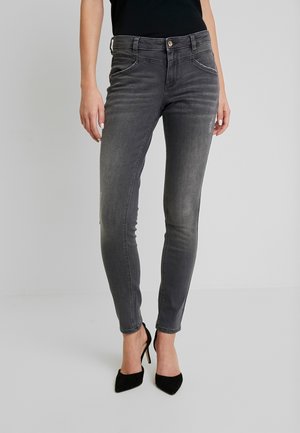 Vaqueros gris ajustados combinados con zapatos negros de tacón alto, mostrando los bolsillos delanteros y laterales en una persona de pie contra un fondo liso.