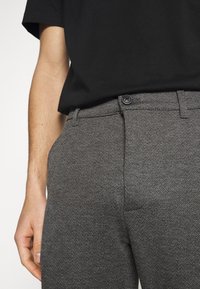 Person wearing gray herringbone-patterned trousers with button closure and a black short-sleeve shirt, hand resting near pocket.