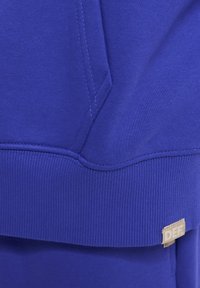 Ljubičasta dukserica s teksturiranim materijalom, rebrastim rukavima i donjim rubom, s malom bež etiketom na kojoj piše "DEF" sprijeda.