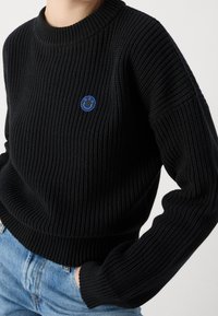 Pull en maille côtelée noire avec un col rond et des manches longues. Présente un petit logo visage souriant bleu sur la poitrine.