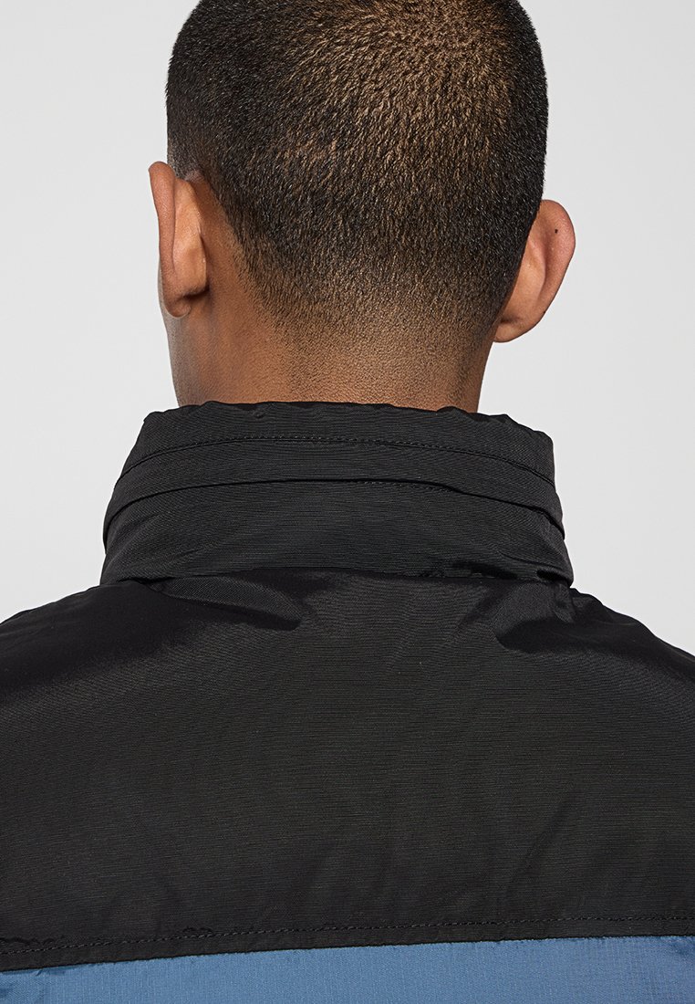Veste noire et bleue avec un col haut, fabriquée en tissu lisse et léger. Présente des blocs de couleur contrastants et des détails de couture subtils.