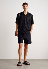 Chemise noire à manches courtes avec un motif texturé, associée à un short cargo noir et des sandales à glissière, portée par un modèle debout.