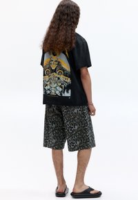 Černé grafické tričko s designem lebky, spárované se zelenými šortkami s leopardím vzorem. Obuv tvoří černé slip-on sandály.
