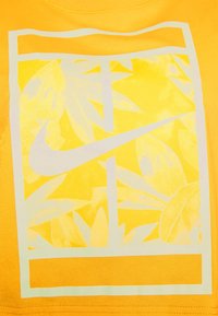Yellow t-shirt featuring a white graphic of overlapping leaves and a Nike logo within a rectangular frame. Soft fabric texture.