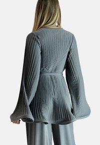 Chaqueta gris plisada con mangas acampanadas largas y cintura ajustada. Textura suave y ligero brillo, con plisados verticales en toda la prenda.