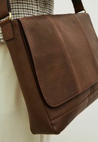 Bolso de hombro de cuero marrón con una textura suave, cierre de solapa y costuras en contraste. Cuenta con una correa cruzada y un diseño minimalista.