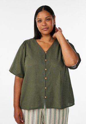 Femme au teint moyen et aux cheveux noirs lisses porte un chemisier vert à manches courtes boutonné et un pantalon clair à rayures verticales.