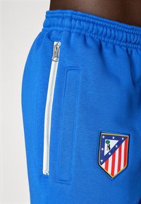 Blue athletic shorts with a textured finish, featuring a white zippered pocket, and a colorful embroidered badge on the side.