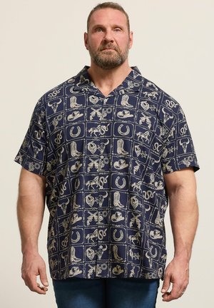 Brodaty mężczyzna noszący granatową koszulę na krótki rękaw zapinaną na guziki, ozdobioną beżowymi, westernowymi motywami, takimi jak buty, podkowy, kapelusze i węże.