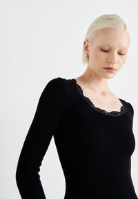 Top preto de manga longa com textura canelada, apresentando um decote com renda. Textura suave com uma silhueta justa.