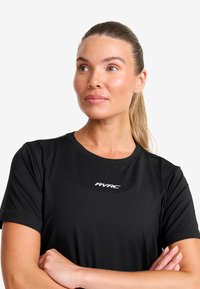 Kvinna med blont hår i en hästsvans, iförd en svart kortärmad tröja med "RVRC"-logotypen, med armarna i kors och tittande lite uppåt mot sidan.