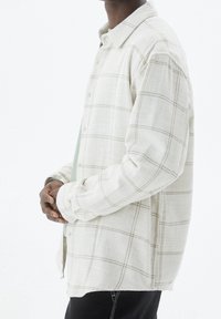 Camisa de cuadros beige claro con cierre de botones, confeccionada en tela texturizada. Tiene mangas largas y se lleva sobre una camiseta verde claro.