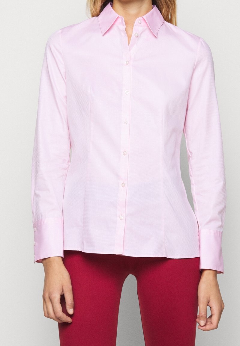Femme portant une chemise rose clair à manches longues boutonnée et un pantalon rouge, debout devant un fond clair uni.