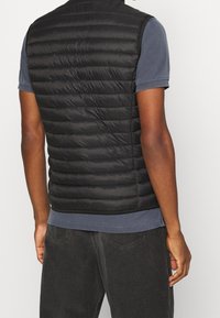 Mann, der eine schwarze gesteppte ärmellose Pufferweste über einem kurzärmeligen navyblauen Poloshirt und einer dunklen Cordhose trägt, von hinten gezeigt.
