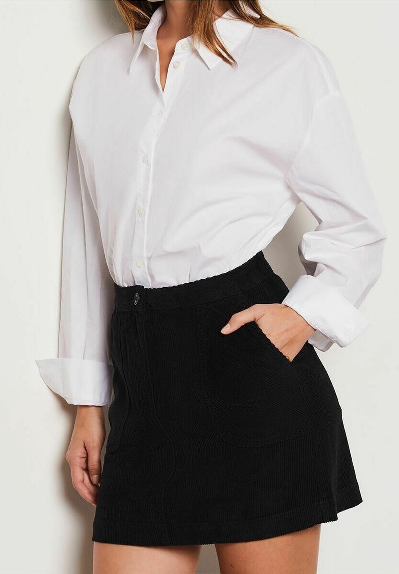 Femme portant une chemise blanche boutonnée rentrée dans une jupe en velours côtelé noire, avec une main dans la poche.