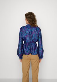 Kobieta o kręconych włosach ma na sobie kolorową bluzkę w fioletowe i niebieskie wzory, w połączeniu z khaki spodniami, widziana z tyłu.