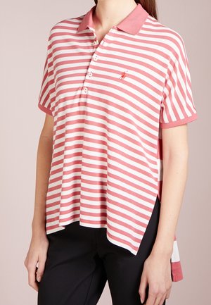 Vrouw in een ruimvallend, korte mouwen roze en witte gestreepte poloshirt met knopen en zwarte broek, staand tegen een effen achtergrond.