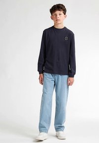 Tmavomodrá košile s dlouhým rukávem s kruhovým logem na hrudi, doplněná světle modrými džínami s volnějším střihem a bílými teniskami.