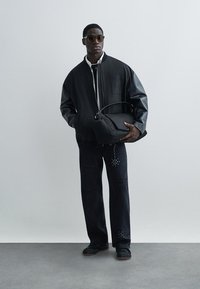 Άνδρας που φοράει μαύρο bomber μπουφάν με δερμάτινα μανίκια, μαύρο τζιν με σχέδια με τρουκς, γυαλιά ηλίου, κρατώντας μια μεγάλη μαύρη τσάντα σε απλό φόντο.