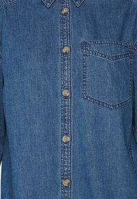 Camicia in denim blu medio con colletto a bottoni, bottoni frontali e taschino sul petto. Presenta una lieve trama e dettagli cuciti.