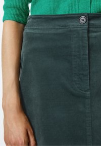 Dunkelblau-grüner Denim-Rock mit glatt strukturiertem Stoff, durchgehender Knopfleiste vorne und Seitentaschen, kombiniert mit einem grünen gerippten Pullover.