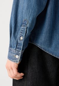 Camicia in denim blu con maniche lunghe, caratterizzata da bottoni decorativi ai polsini, cuciture a vista e un orlo leggermente curvo.