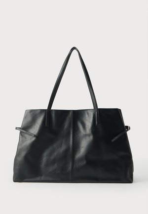 Grand sac cabas en cuir noir avec double bandoulière et détails noués sur les côtés, présenté sur un fond clair uni.