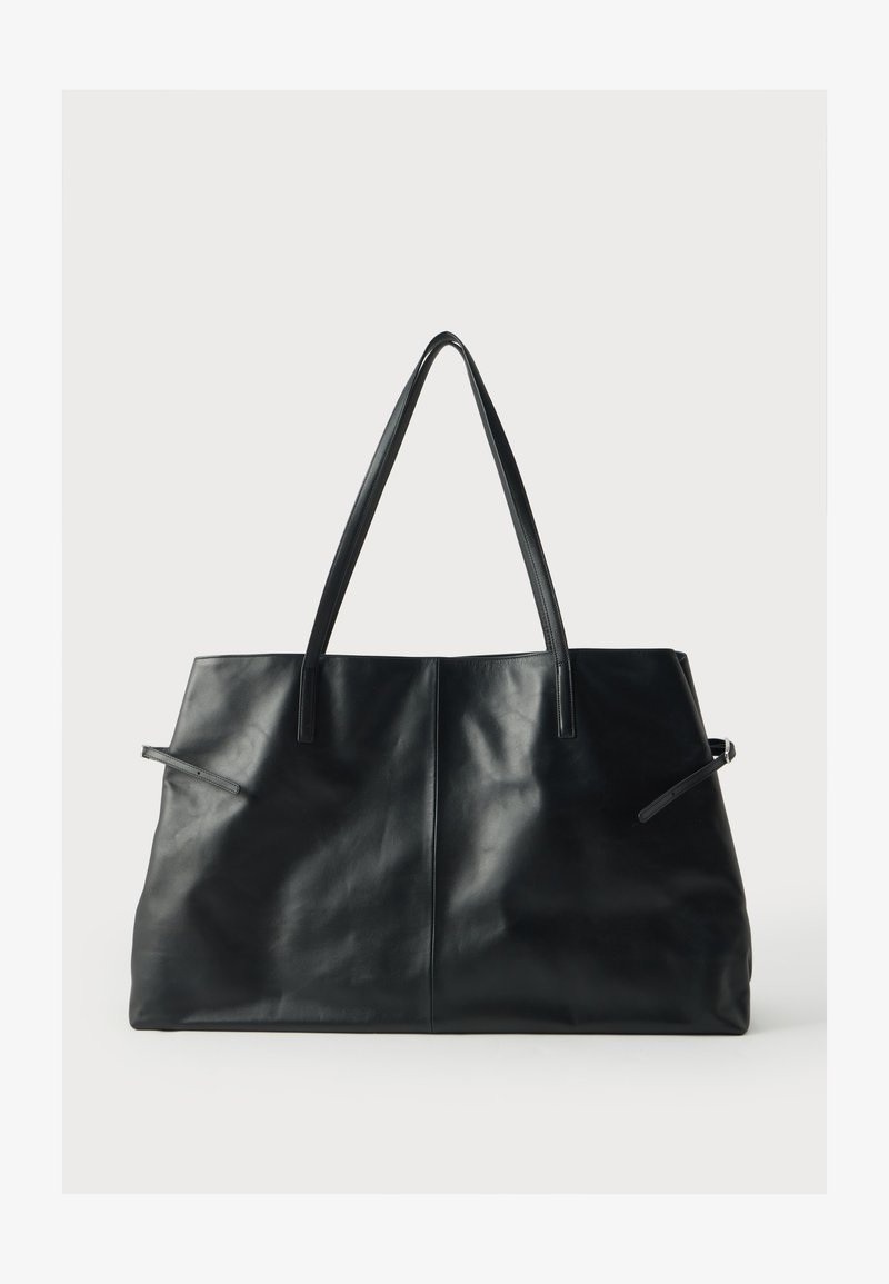 Grand sac cabas en cuir noir avec double bandoulière et détails noués sur les côtés, présenté sur un fond clair uni.