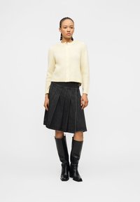 Γυναίκα που φοράει κρεμ πλεκτή ζακέτα, μαύρη πλισέ φούστα από denim και μαύρα δερμάτινα μπότες μέχρι το γόνατο, στέκεται μπροστά από μια απλή φόντο.