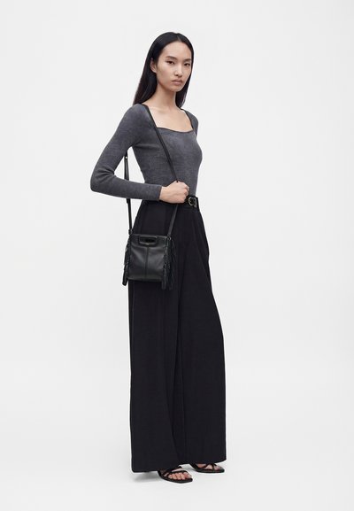 Femme aux longs cheveux noirs portant un haut gris à manches longues, un pantalon large noir, des sandales noires et un sac bandoulière noir avec des détails à franges.
