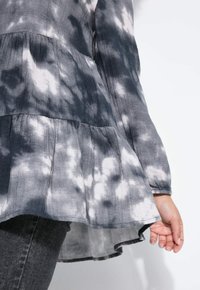 Grijs tie-dye blouse met een gelaagd peplum ontwerp. Heeft lange mouwen, een gebogen zoom en een lichte stoftextuur.