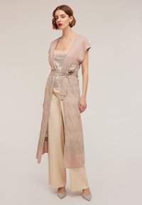 Long, beige outer garment with short sleeves, textured weave, a tie belt at the waist, layered over a sequined top and cream trousers.